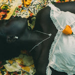 A close-up lifestyle shot of a person lying down wearing a white lace-trimmed top, with a small orange flower pinned to the chest, against a colorful patterned textile background.