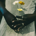 A close-up of a person in a white lace-trimmed dress holding three small wildflowers between two crossed dark hands with rings and painted fingernails.