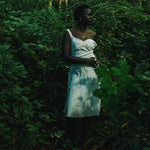 A woman stands in a dense green forest wearing a silver necklace and a white sleeveless dress, surrounded by lush foliage and trees.