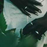 A close-up lifestyle shot of a person's hands with silver rings, resting on a white fabric surface with green leaves in the foreground.