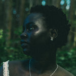 A woman in a white lace-trim top wearing a delicate silver pendant necklace and matching silver stud dangle earrings, photographed outdoors in a dark, green-toned forest setting.