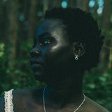 A woman in a white lace-trim top wearing a delicate silver pendant necklace and matching silver stud dangle earrings, photographed outdoors in a dark, green-toned forest setting.