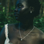 A woman in a white lace-trim top wearing a delicate silver pendant necklace and matching silver hoop earrings, photographed outdoors in a dark, green-toned forest setting.