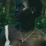 A woman in a white lace-trim top wearing a delicate silver pendant necklace and matching silver hoop earrings, photographed outdoors in a dark, green-toned forest setting.