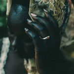 A close-up portrait of a person wearing a patterned headscarf in earthy tones and a silver ring with a small orange stone, with their hand resting against their face.