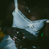 A close-up lifestyle shot of a person wearing a white lace-trimmed camisole top and white pants, and silver rings visible against a very dark background.