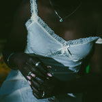 A close-up lifestyle shot of a person wearing a white lace-trimmed camisole top and white pants, adorned with silver rings.