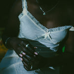 A close-up lifestyle shot of a person wearing a white lace-trimmed camisole top and white pants, and silver rings visible against a very dark background.