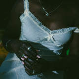A close-up lifestyle shot of a person wearing a white lace-trimmed camisole top and white pants, and silver rings visible against a very dark background.