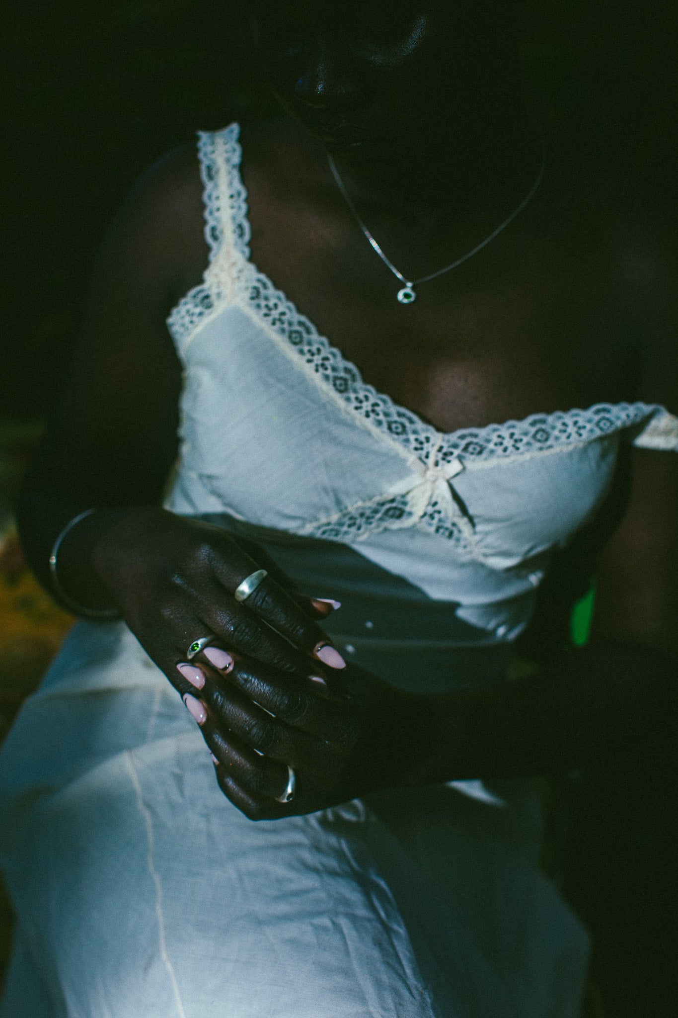 A close-up lifestyle shot of a person wearing a white lace-trimmed camisole top and white pants, and silver rings visible against a very dark background.