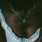 A close-up lifestyle shot of a person wearing a delicate silver chain necklace with a small round pendant, styled with a white lace-trimmed top.