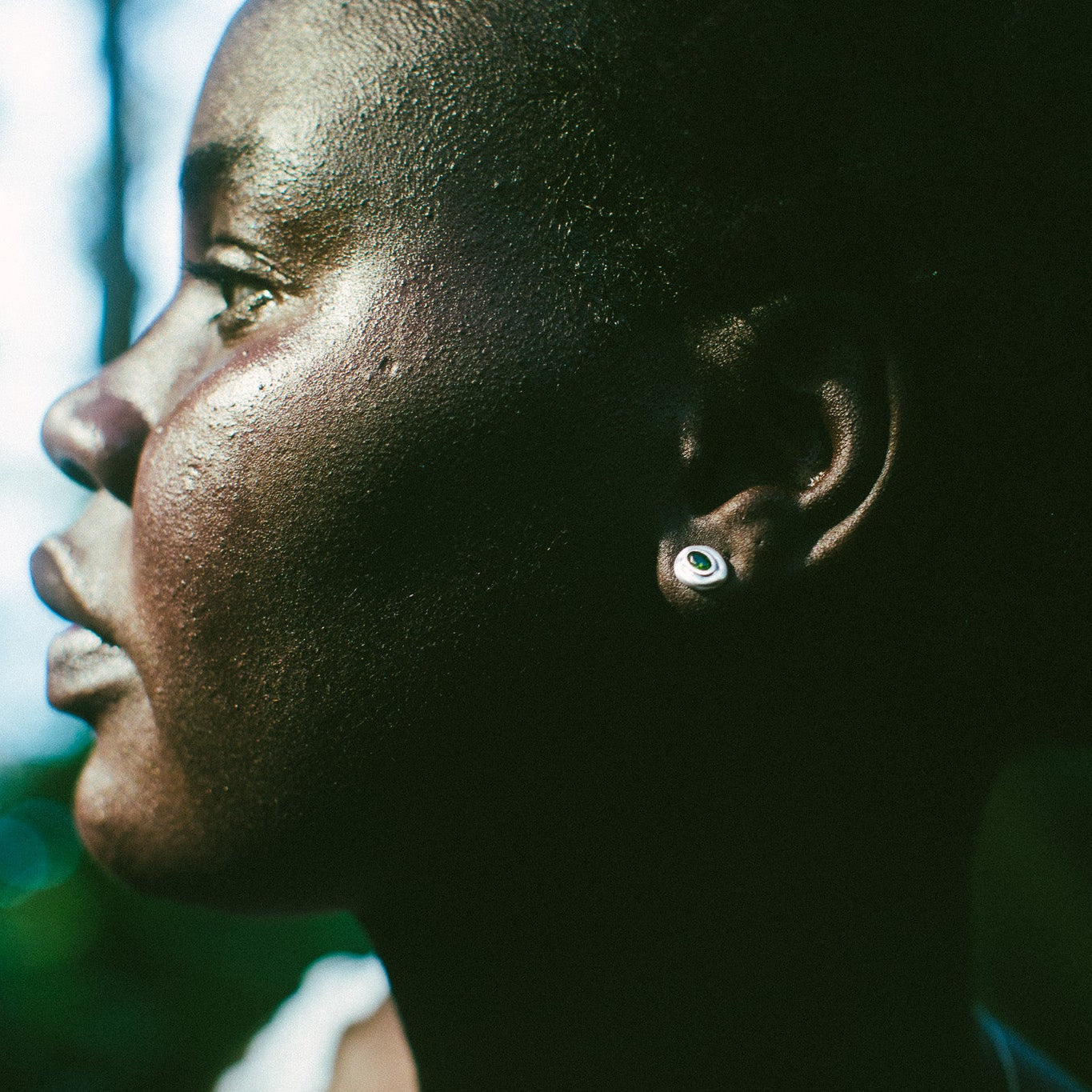 Close-up side profile of a person outdoors wearing a small round earring with a green center.