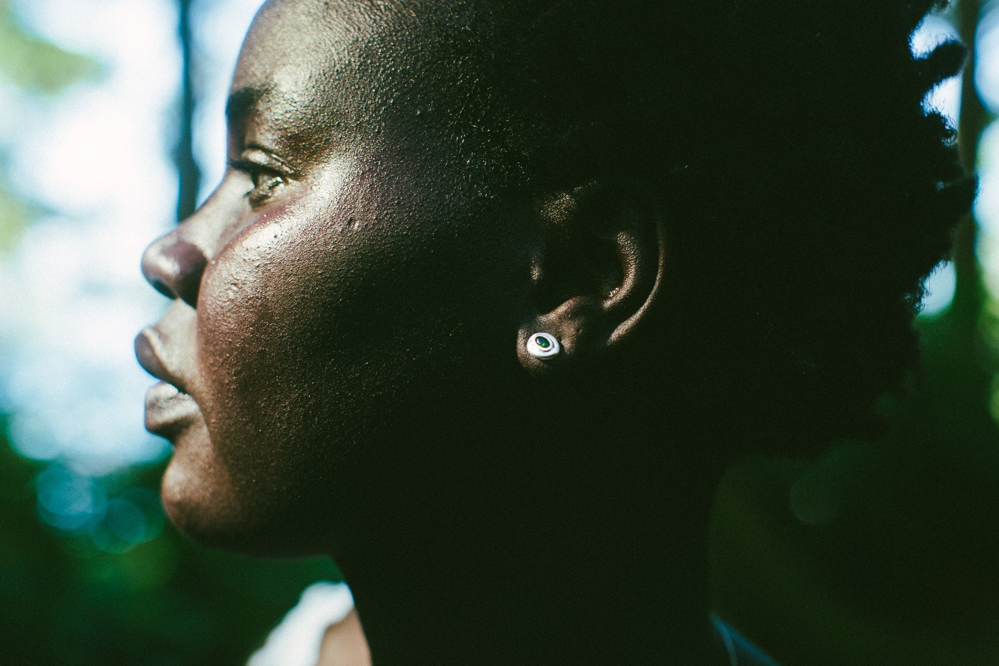Close-up side profile of a person outdoors wearing a small round earring with a green center.