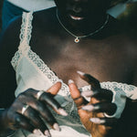 A close-up lifestyle shot of a person wearing a white lace-trimmed camisole top, showing their hands with long manicured nails and rings.