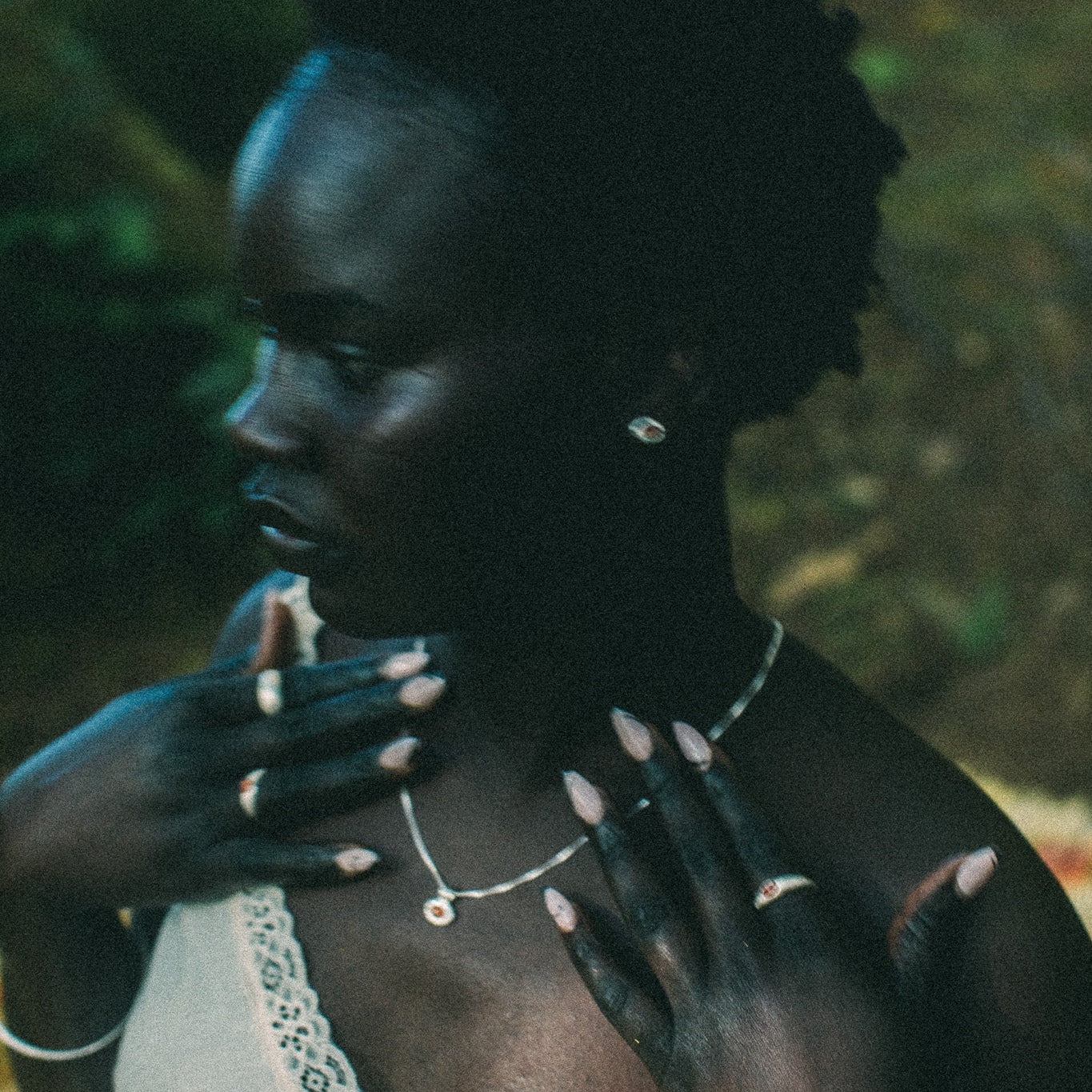 A woman in profile wearing a delicate silver necklace and rings, with her hands near her chest, set against a dark, blurred outdoor background.