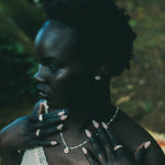 A woman in profile wearing a delicate silver necklace and rings, with her hands near her chest, set against a dark, blurred outdoor background.