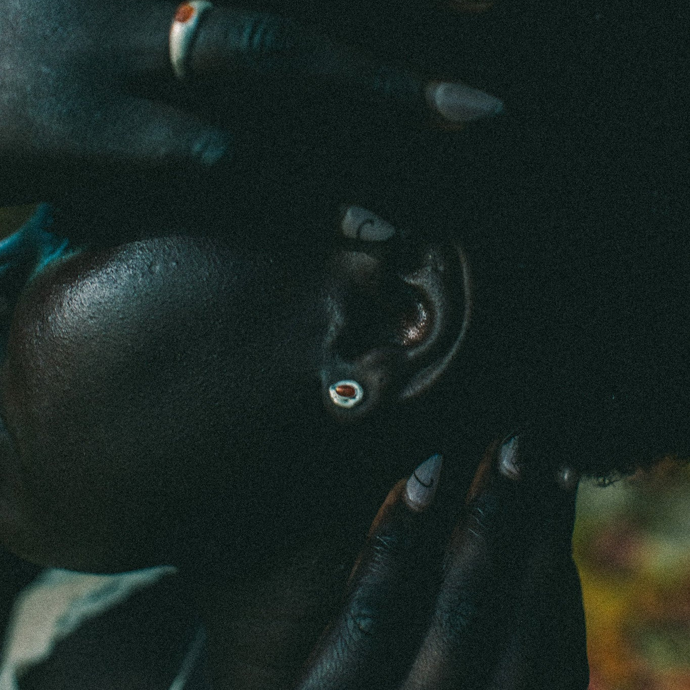 A close-up lifestyle shot of a person with dark skin and short black hair, wearing silver earrings and rings, with their hands resting on their head against a dark, blurred background.