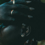 A close-up lifestyle shot of a person wearing silver-toned earrings and rings, with their hands resting on their head against a dark, blurred background.