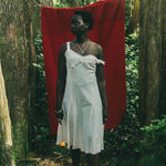 A woman stands barefoot in a forest wearing a white one-shoulder dress, with a large red cloth hanging behind her between tree trunks.