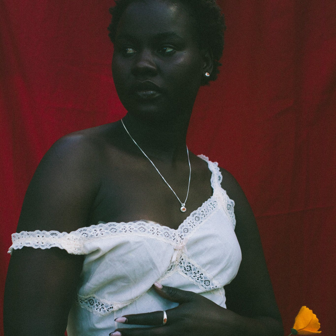 A woman stands against a red fabric backdrop wearing a white one-shoulder dress with lace trim and holding a small yellow flower.