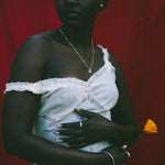 A woman stands against a red fabric backdrop wearing a white one-shoulder dress with lace trim and holding a small yellow flower.