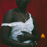 A woman stands against a red fabric backdrop wearing a white one-shoulder dress with lace trim and holding a small yellow flower.