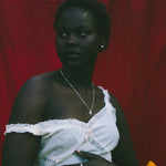 A woman stands against a red fabric backdrop wearing a white one-shoulder dress with lace trim and holding a small yellow flower.