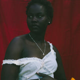A woman stands against a red fabric backdrop wearing a white one-shoulder dress with lace trim and holding a small yellow flower.