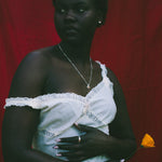 A woman stands against a red fabric backdrop wearing a white one-shoulder dress with lace trim and holding a small yellow flower, wearing a silver necklace and silver ring with orange gemstones.