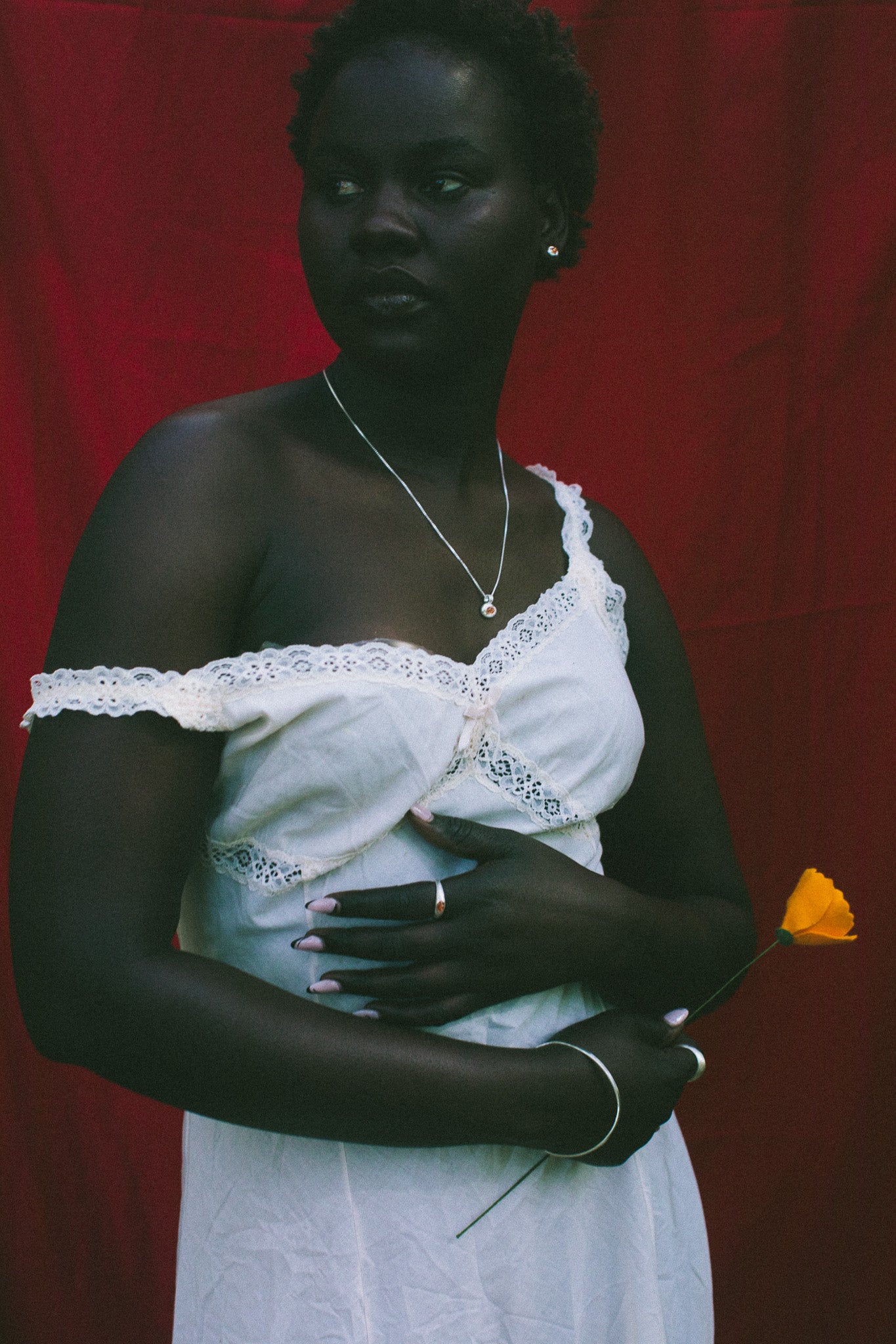 A woman stands against a red fabric backdrop wearing a white one-shoulder dress with lace trim and holding a small yellow flower, wearing a silver necklace and silver ring with orange gemstones.