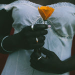 A close-up of a person wearing a white lace-trimmed dress, holding a single orange flower. The person is also wearing several silver rings and a delicate necklace.