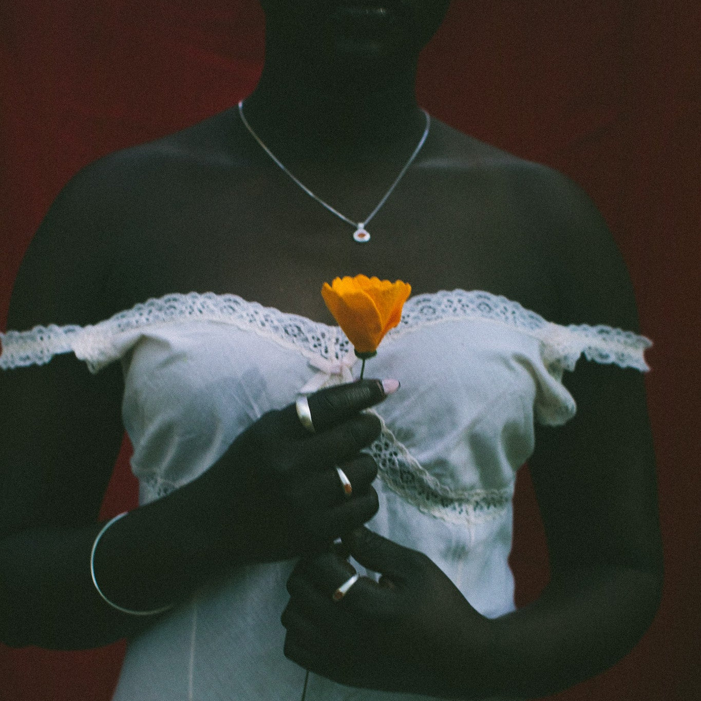 A person wearing a white off-the-shoulder lace-trim dress stands against a dark red background, holding a single orange flower. The person is also wearing a silver necklace and rings.