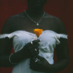 A person wearing a white off-the-shoulder lace-trim dress stands against a dark red background, holding a single orange flower. The person is also wearing a silver necklace and rings.