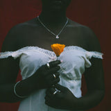 A person wearing a white off-the-shoulder lace-trim dress stands against a dark red background, holding a single orange flower. The person is also wearing a silver necklace and rings.