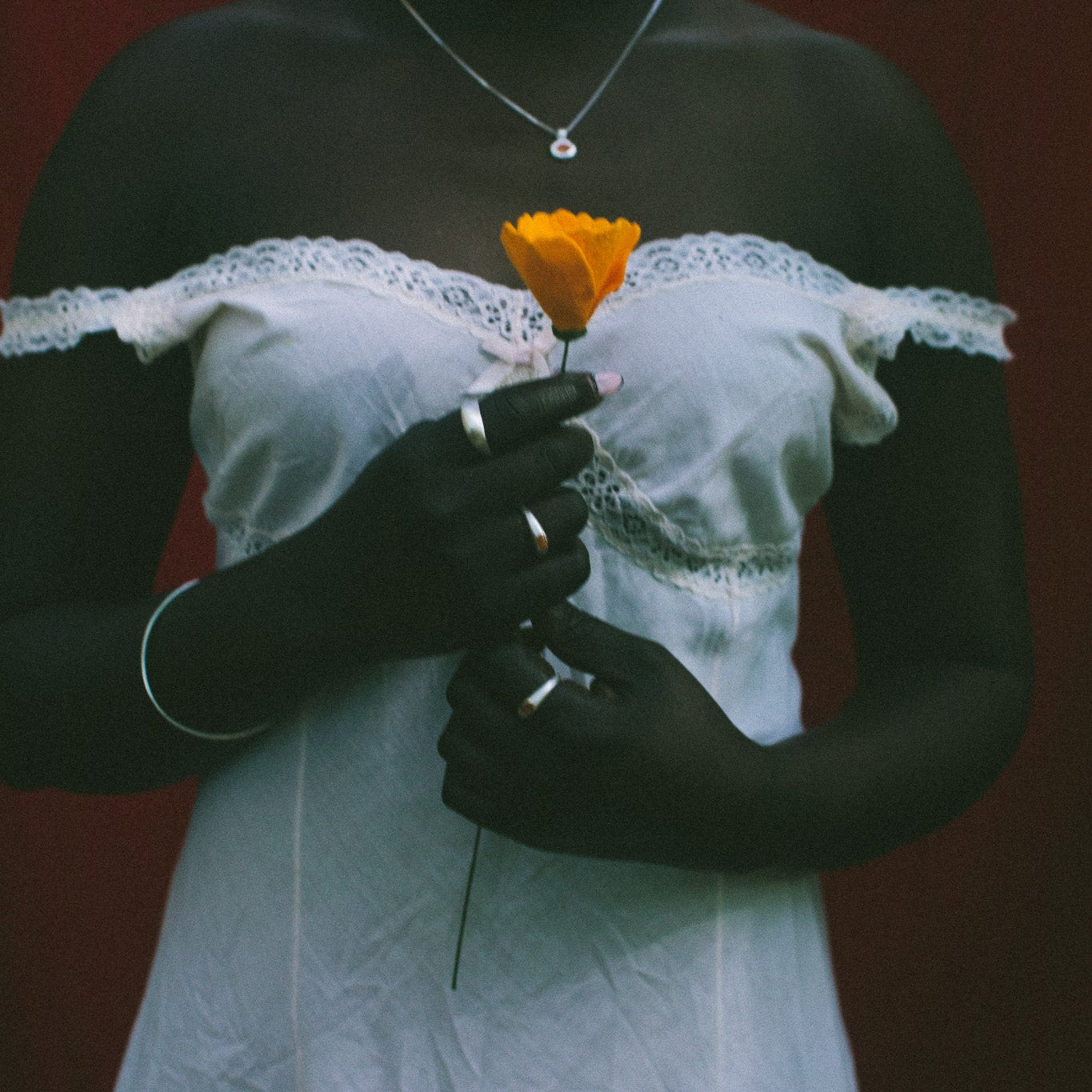 A person wearing a white off-the-shoulder lace-trim dress stands against a dark red background, holding a single orange flower. The person is also wearing a silver necklace and rings.
