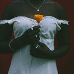 A person wearing a white off-the-shoulder lace-trim dress stands against a dark red background, holding a single orange flower. The person is also wearing a silver necklace and rings.