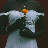 A person wearing a white off-the-shoulder lace-trim dress stands against a dark red background, holding a single orange flower. The person is also wearing a silver necklace and rings.
