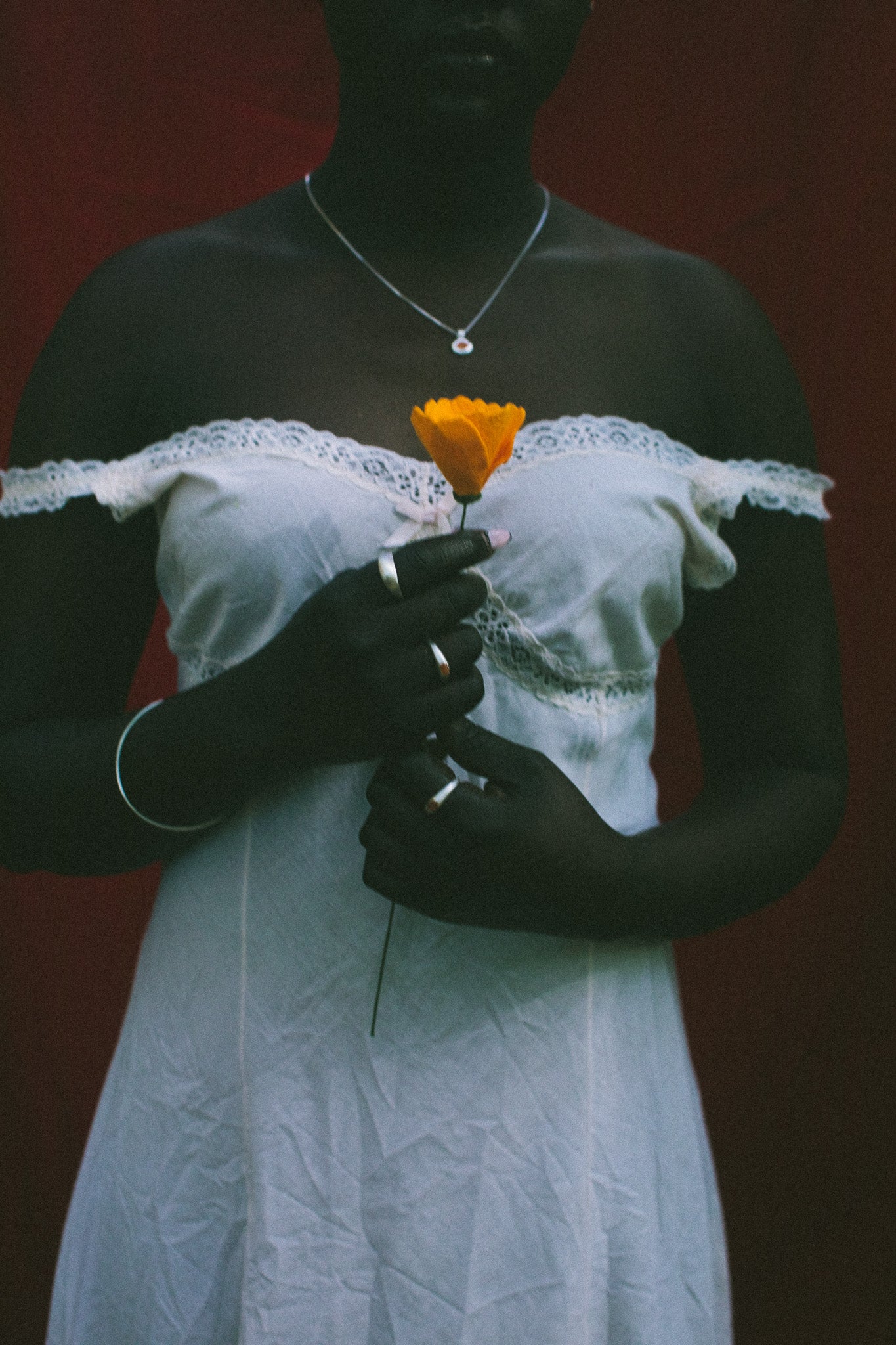 A person wearing a white off-the-shoulder lace-trim dress stands against a dark red background, holding a single orange flower. The person is also wearing a silver necklace and rings.