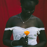 A woman wearing a white off-the-shoulder lace dress, holding a bright orange flower in front of a red draped backdrop.