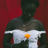 A woman wearing a white off-the-shoulder lace dress, holding a bright orange flower in front of a red draped backdrop.