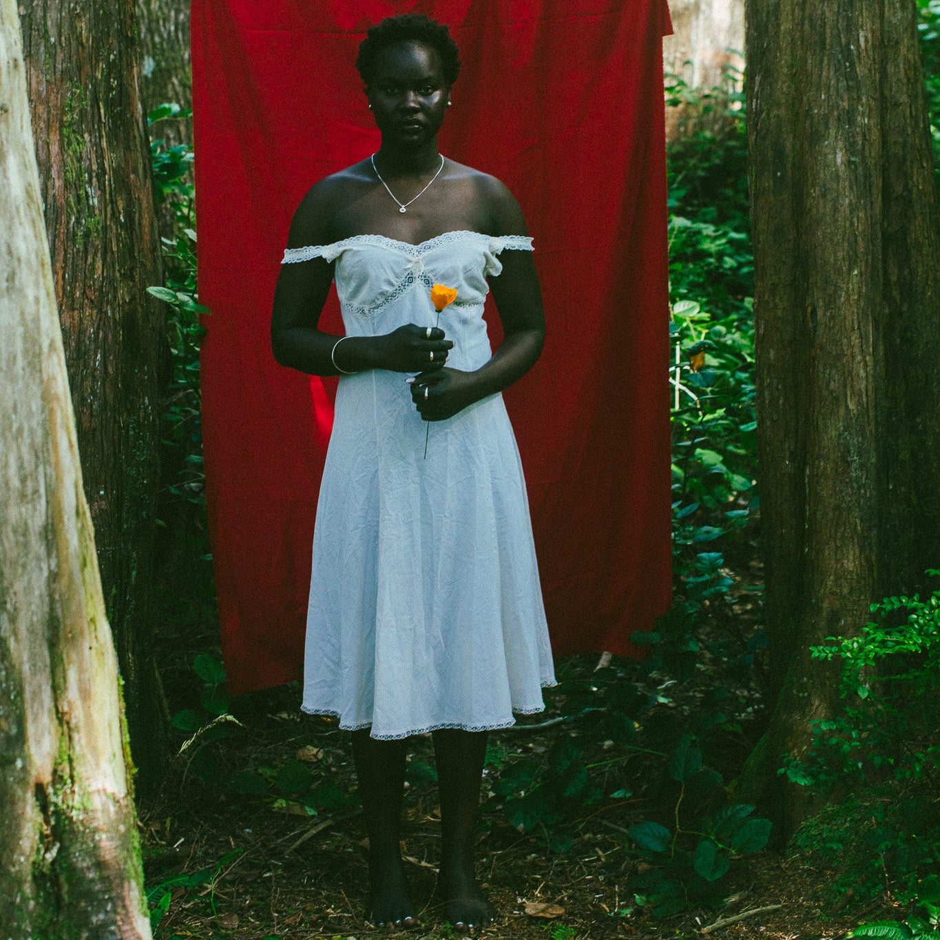 A woman stands in a forest wearing a white off-the-shoulder dress, holding a small orange flower in front of a large red fabric backdrop strung between trees.