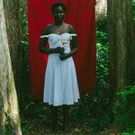 A woman stands in a forest wearing a white off-the-shoulder dress, holding a small orange flower in front of a large red fabric backdrop strung between trees.