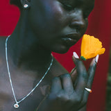 A close-up portrait of a woman holding a bright yellow flower near her face, wearing a silver necklace and earring against a vivid red background.