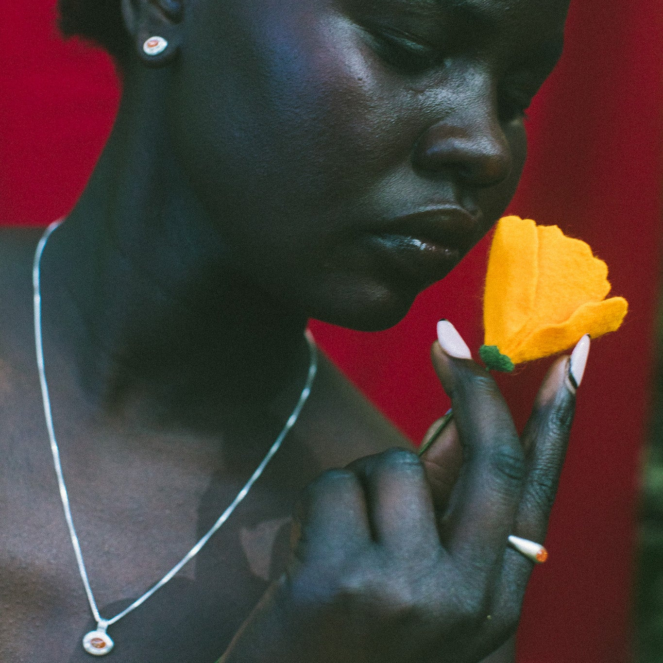 A close-up portrait of a woman holding a bright yellow flower near her face, wearing a silver necklace and earring against a vivid red background.