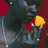 A close-up portrait of a woman holding a bright yellow flower near her face, wearing a silver necklace and earring against a vivid red background.