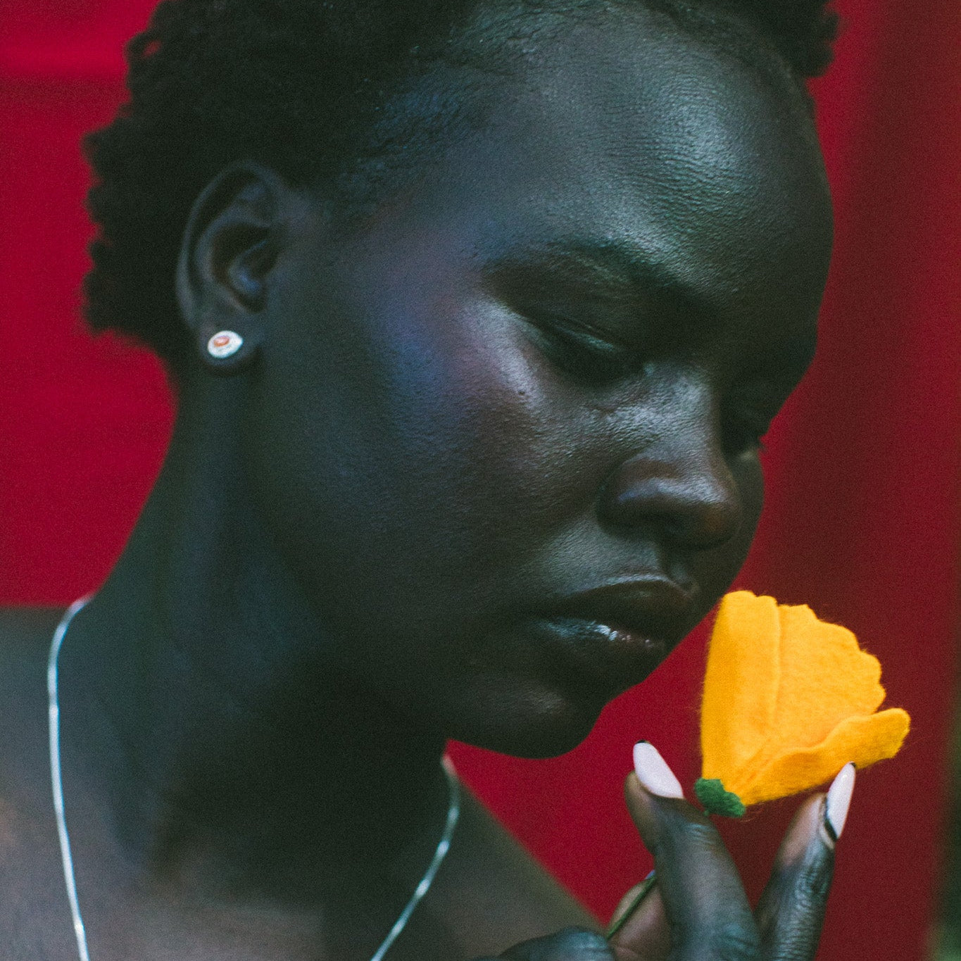 A close-up portrait of a woman holding a bright yellow flower near her face, wearing a silver necklace and earring against a vivid red background.
