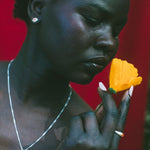 A close-up portrait of a woman holding a bright yellow flower near her face, wearing a silver necklace and earring against a vivid red background.