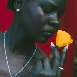 A close-up portrait of a woman holding a bright yellow flower near her face, wearing a silver necklace and earring against a vivid red background.