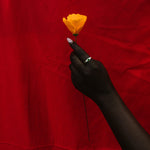 A hand wearing a silver ring holds a single yellow flower against a draped red fabric background.