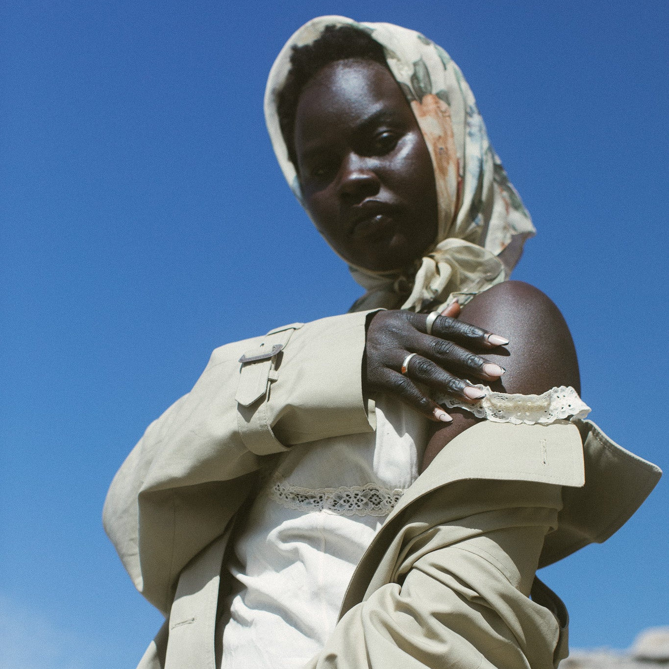 A model wearing a beige trench coat over a white top and trousers, paired with a patterned beige and orange headscarf, set against a bright blue sky and rocky background.